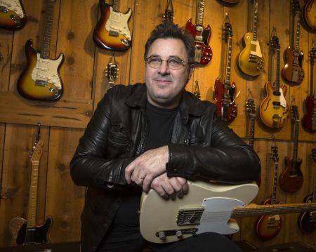 Vince Gill in a leather jacket poses a picture at a guitar shop.